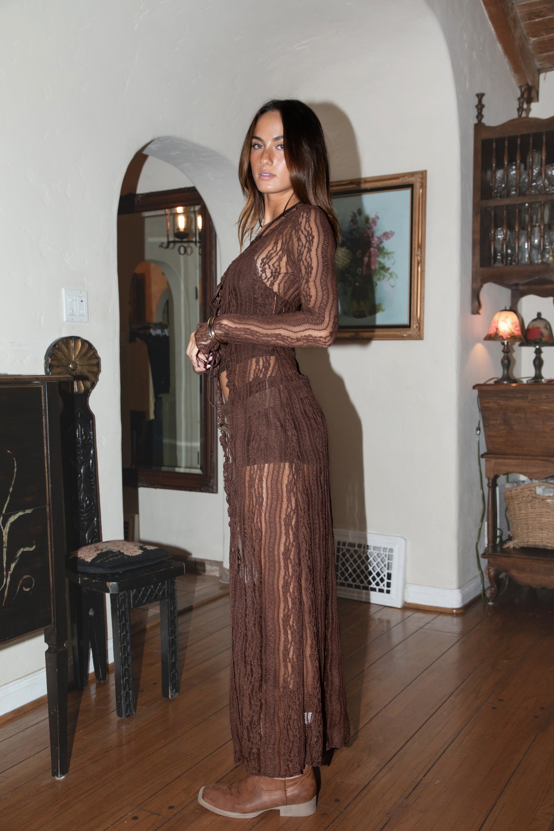 Woman in a brown lace outfit standing in a room with wooden floors and a mirror.