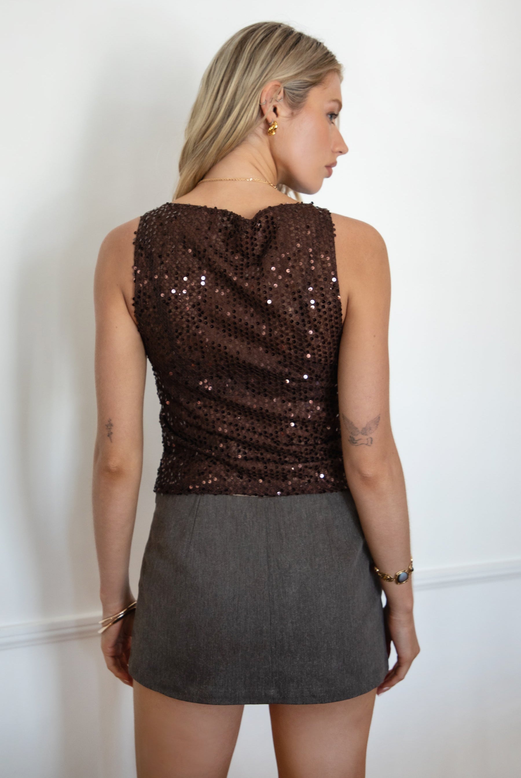 Model wearing the TIC TOC TC31578 LIIMA brown sheer sequin tank top featuring a high boat neckline, sleeveless silhouette, and shimmering mesh fabric paired with a brown denim mini skirt.