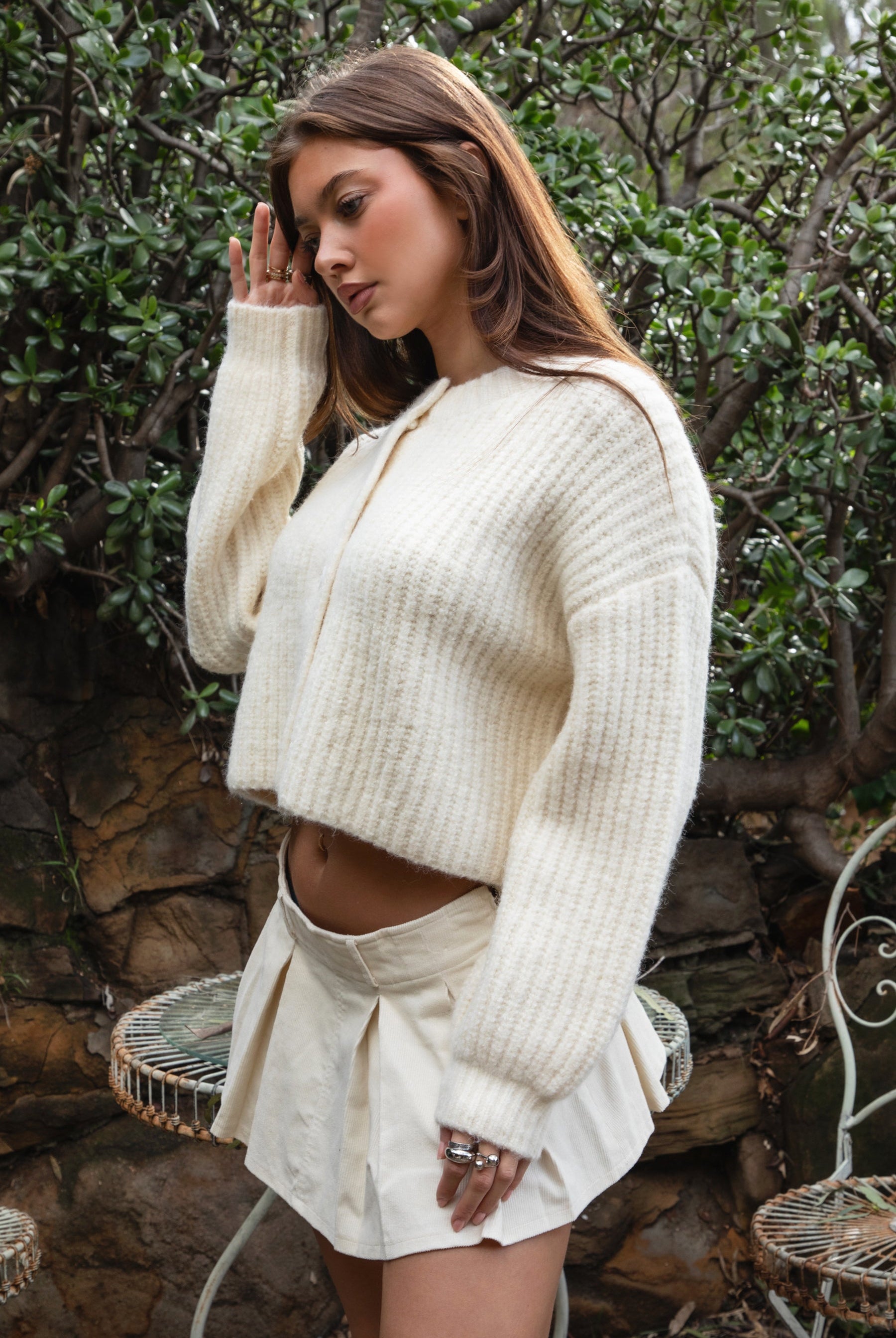 Woman wearing TIC TOC TF31833 ISOLDE cream wool-blend button down cardigan with boxy cropped fit, ribbed trim, and marled knit texture paired with a skirt outdoors with greenery in the background