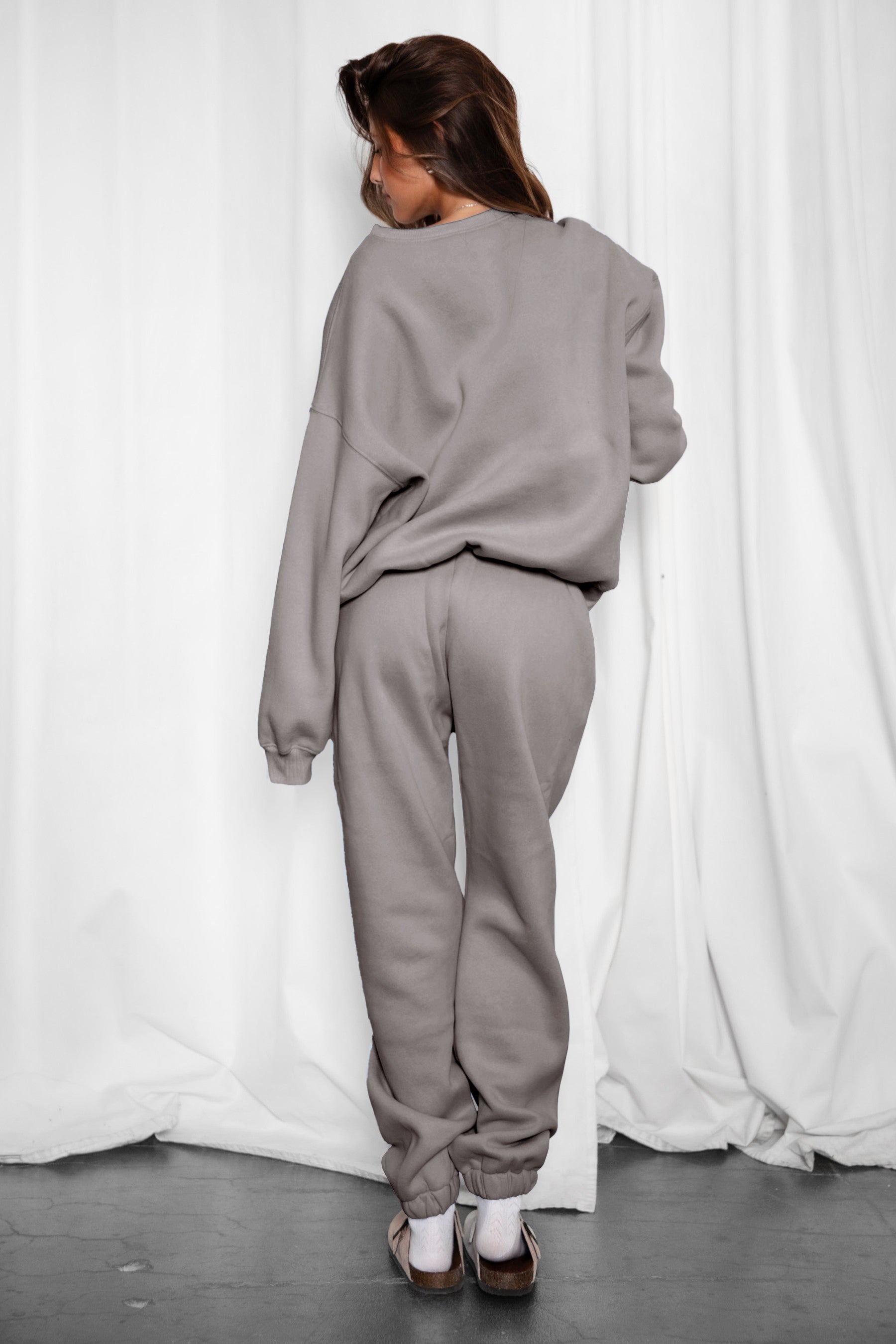 Person wearing a gray sweatshirt and pants against a white curtain background