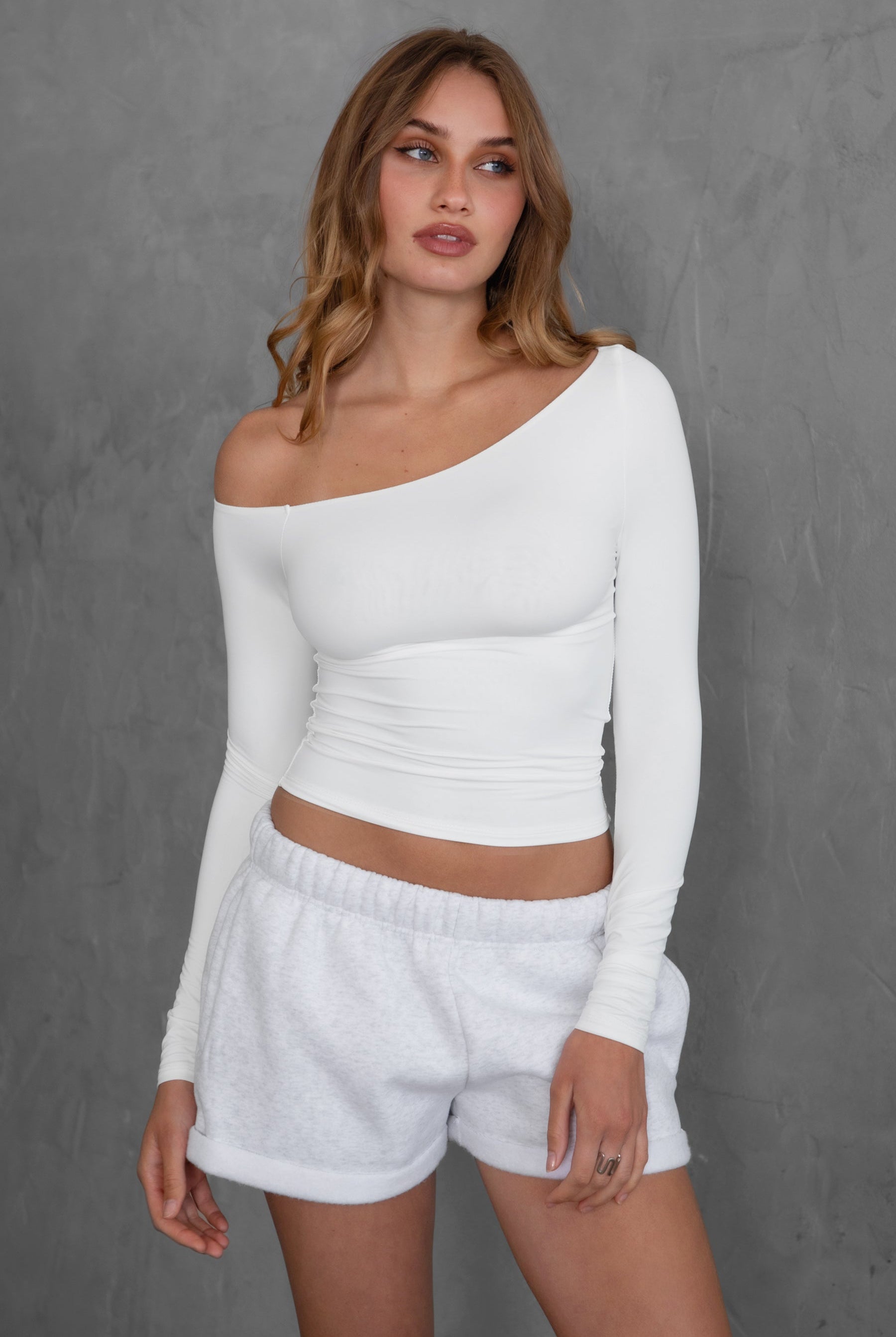 Woman wearing TIC TOC TH30538 AUDREE lightweight one-shoulder long sleeve top in slim fit stretch fabric in color white paired with fleece shorts.