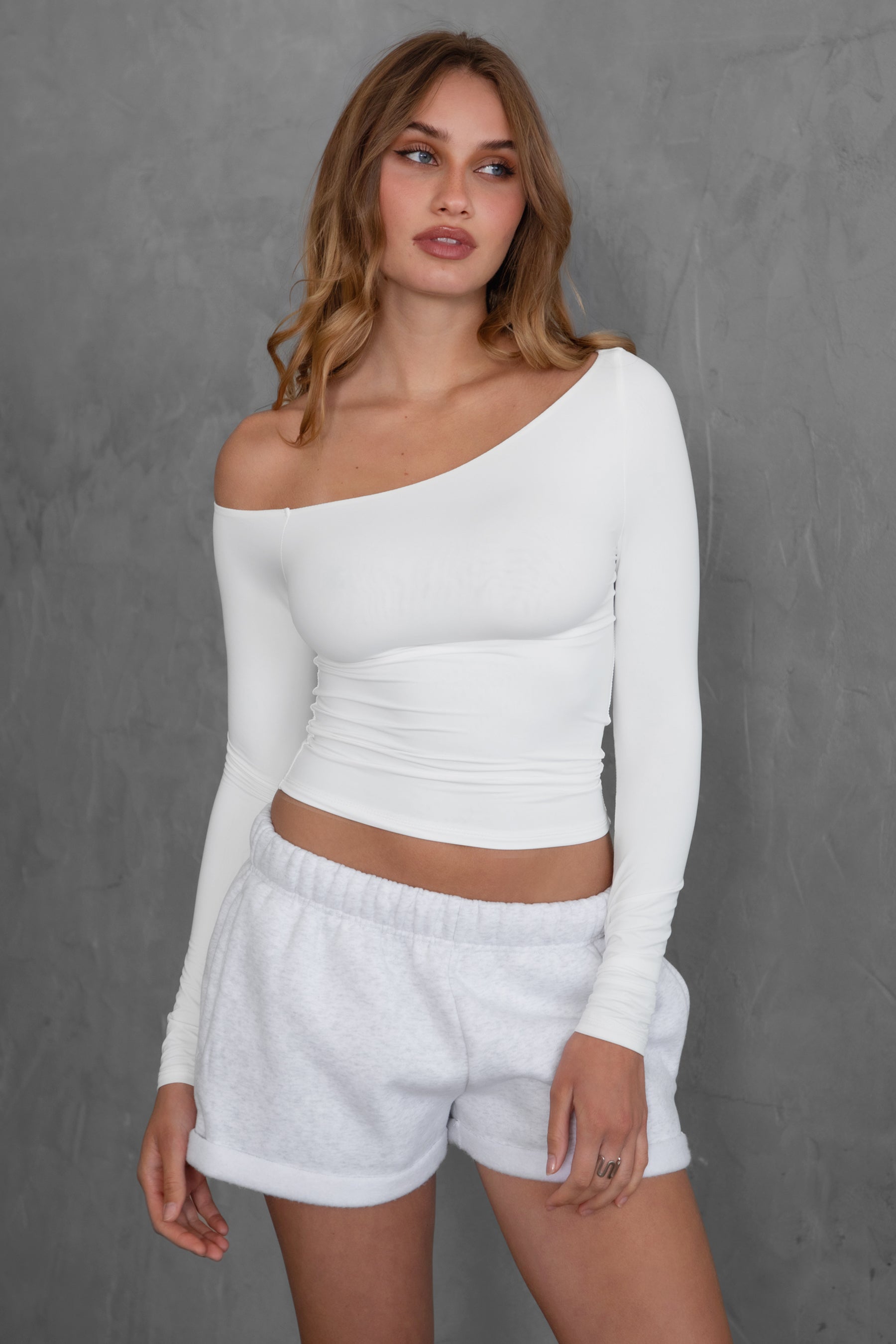 Woman wearing TIC TOC TH30538 AUDREE lightweight one-shoulder long sleeve top in slim fit stretch fabric in color white paired with fleece shorts.