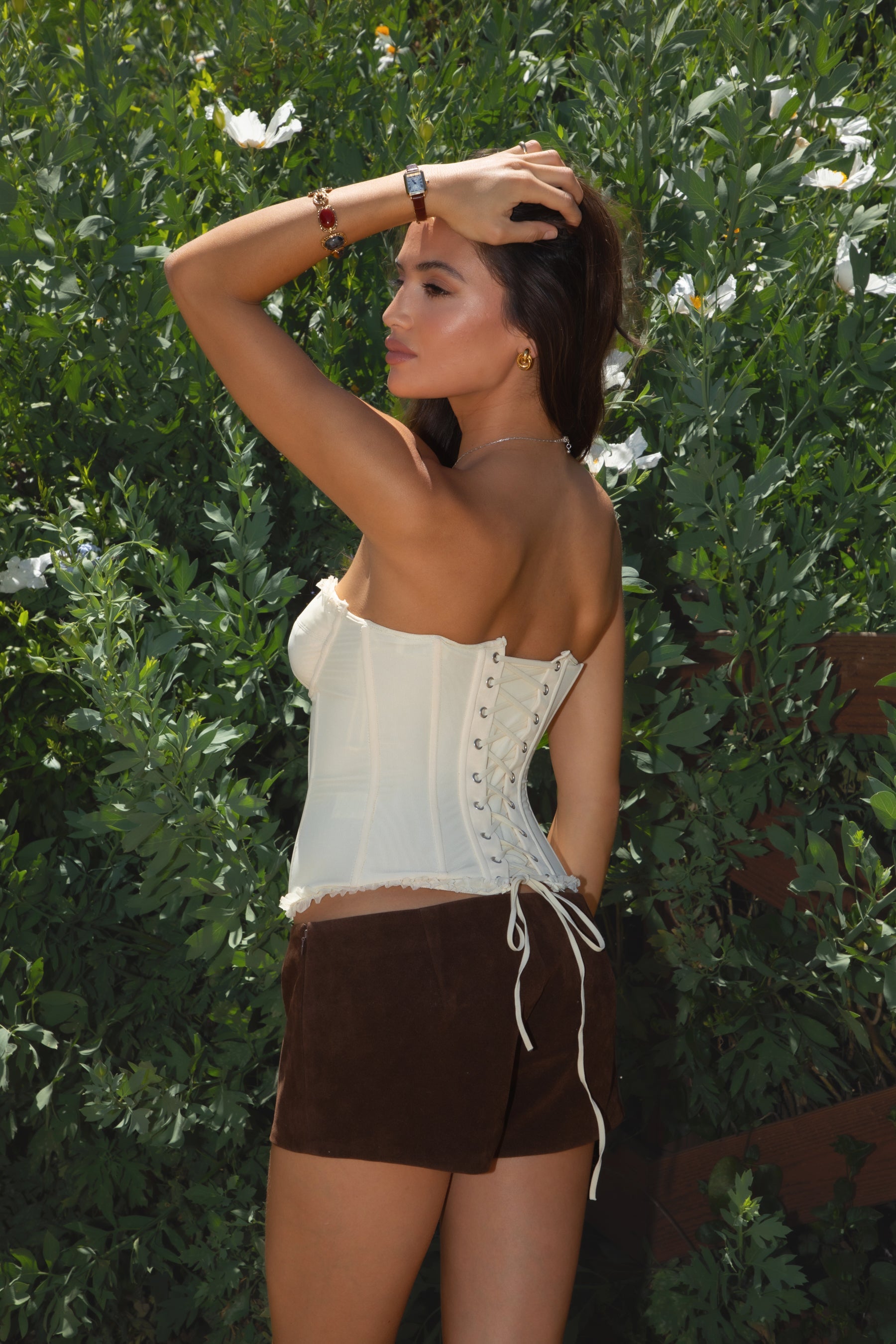 Model wearing the TIC TOC TH30975 LAUREL cream mesh strapless bustier corset top featuring ruffle trim edges, underwire cups, and lace-up back paired with brown suede shorts, standing against greenery