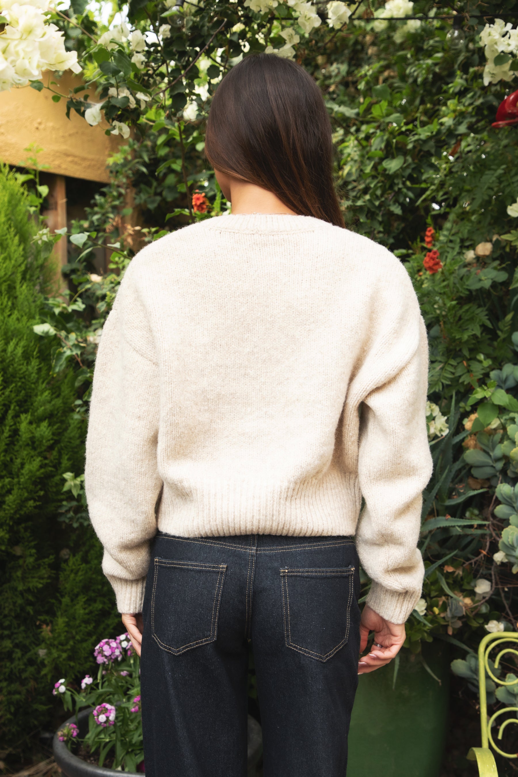 Woman wearing TIC TOC TV31637 MARLO oatmeal sweater knit button down cardigan with ribbed cuffs and dropped shoulders in cozy marled fabric paired with dark denim in a garden setting