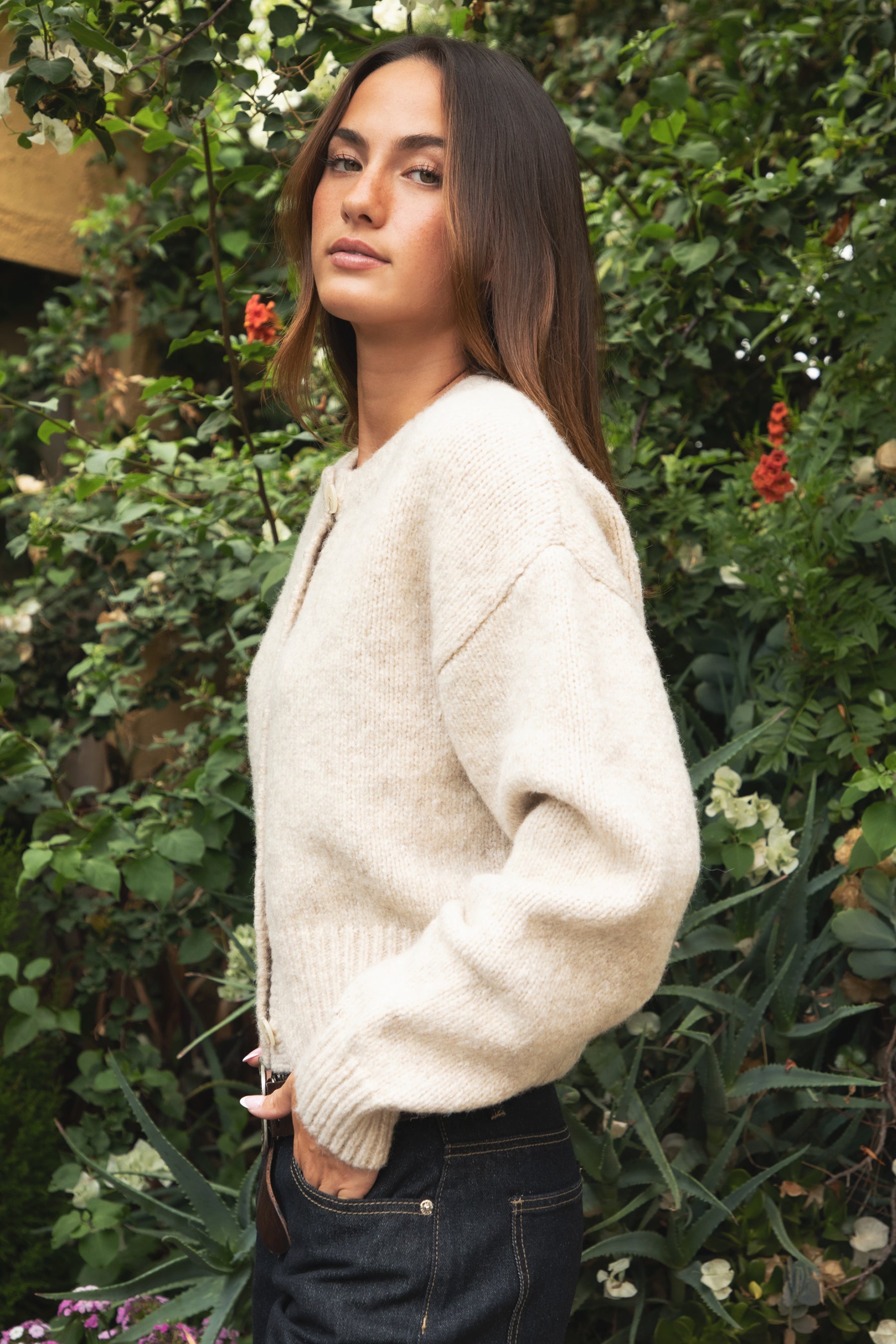 Woman wearing TIC TOC TV31637 MARLO oatmeal sweater knit button down cardigan with ribbed cuffs and dropped shoulders in cozy marled fabric paired with dark denim in a garden setting