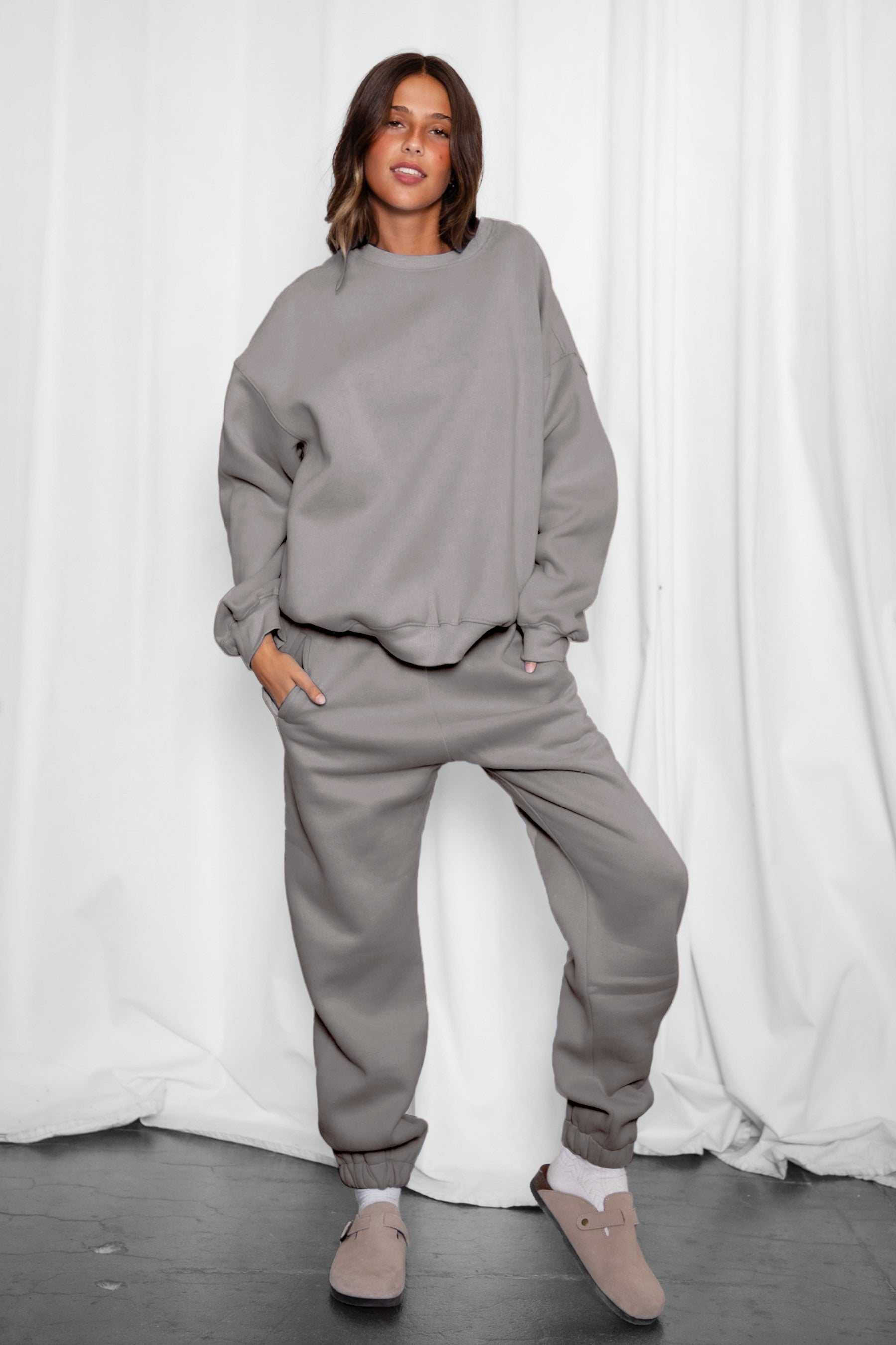 Person wearing a gray sweatshirt and pants set against a white curtain background