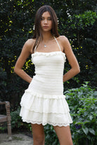 Woman wearing TIC TOC DA32031 ELLIE smocked body tiered ruffle lace halter mini dress featuring embroidered poplin fabric, a sculpted smocked bodice, halter neckline with self-tie strap, drop waist silhouette, and a tiered mini skirt with double layer eyelet lace hem for a romantic summer occasion look.