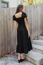 Woman wearing WYNORA smocked puff sleeve tiered midi dress featuring smooth stretch poplin fabric, soft jersey lining, square neckline with adjustable front tie detail, smocked bodice, short puff sleeves with ribbon tie accents, and a flowing tiered midi skirt with ruffle hem finish.