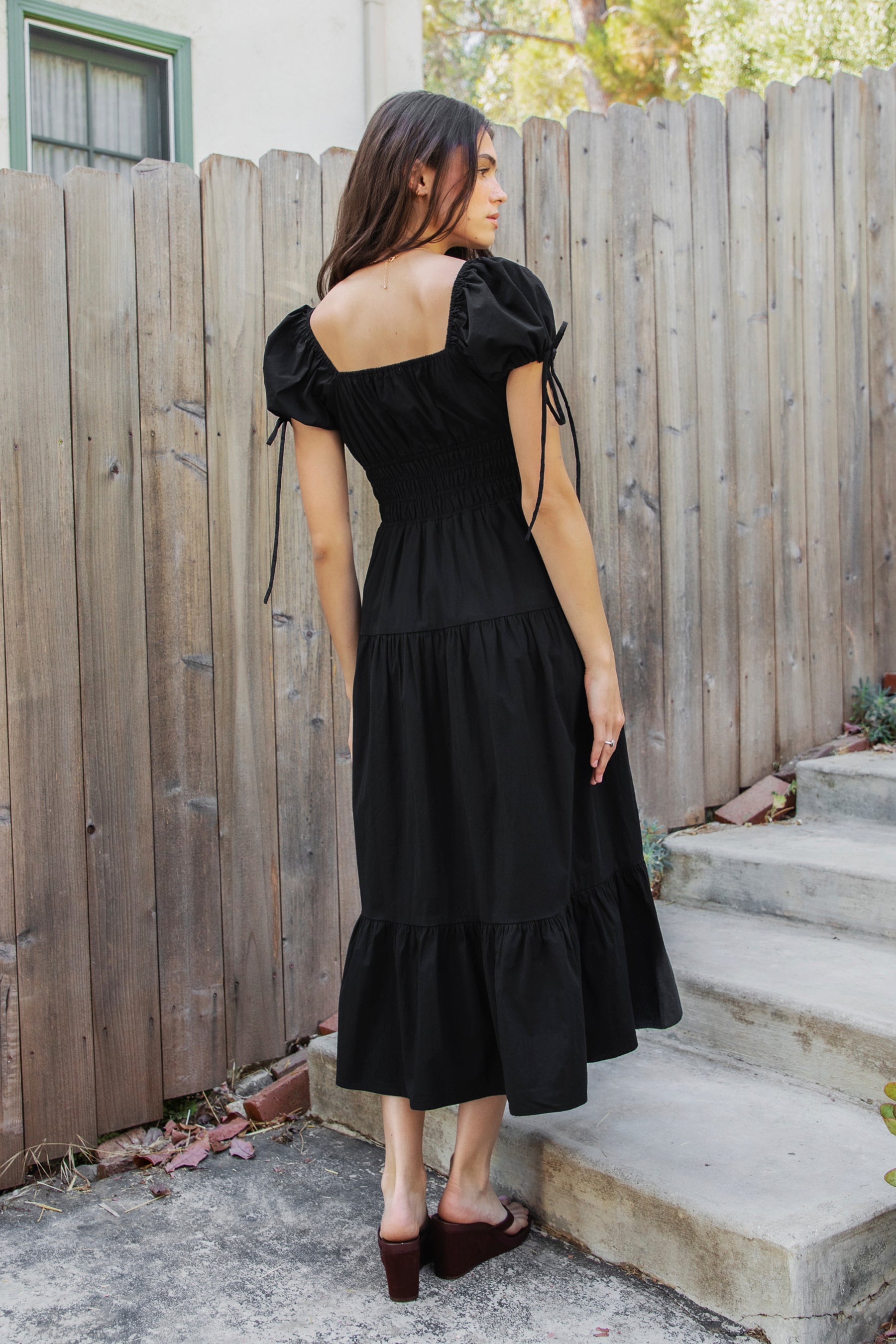 Woman wearing WYNORA smocked puff sleeve tiered midi dress featuring smooth stretch poplin fabric, soft jersey lining, square neckline with adjustable front tie detail, smocked bodice, short puff sleeves with ribbon tie accents, and a flowing tiered midi skirt with ruffle hem finish.