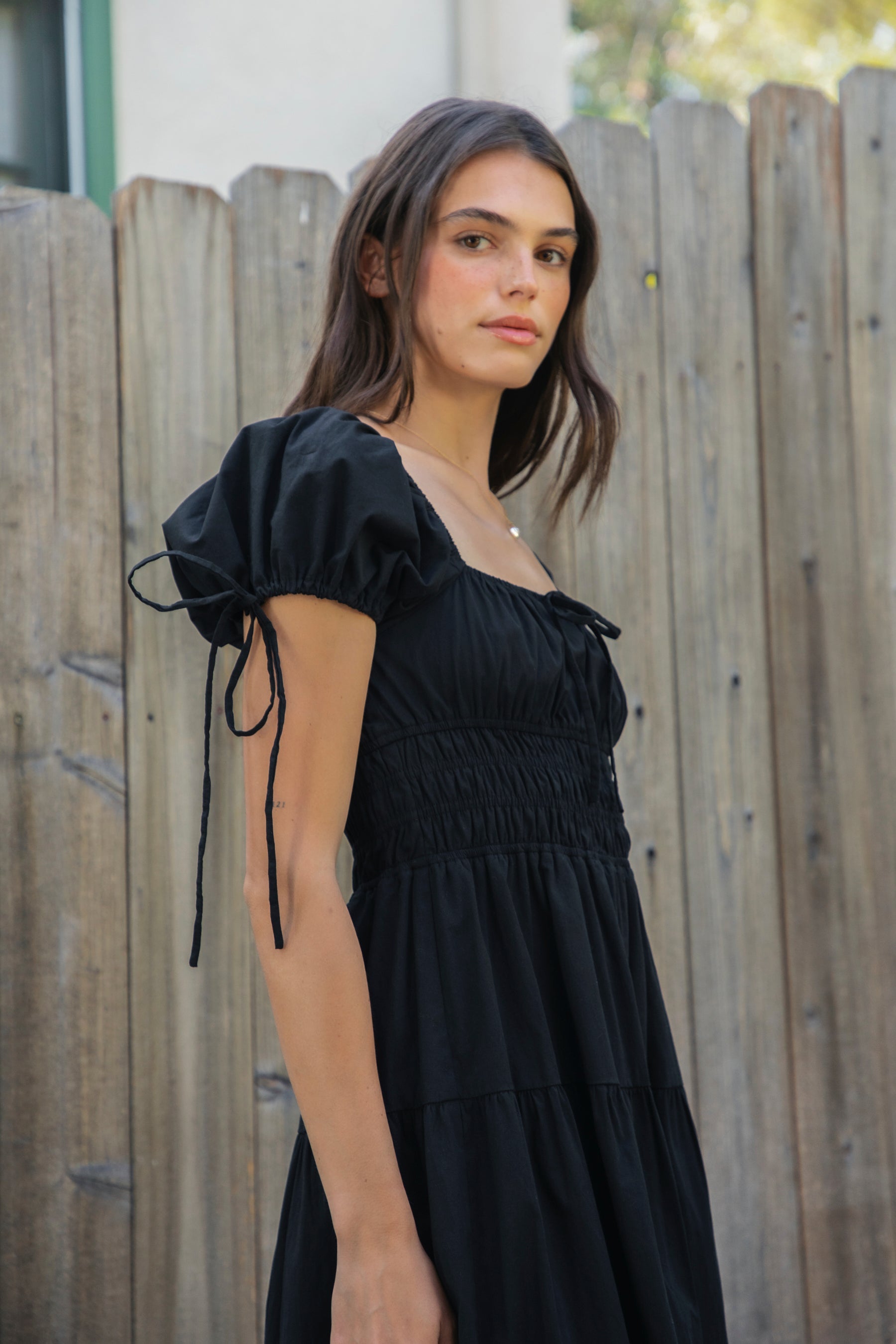 Woman wearing WYNORA smocked puff sleeve tiered midi dress featuring smooth stretch poplin fabric, soft jersey lining, square neckline with adjustable front tie detail, smocked bodice, short puff sleeves with ribbon tie accents, and a flowing tiered midi skirt with ruffle hem finish.