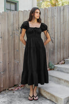 Woman wearing WYNORA smocked puff sleeve tiered midi dress featuring smooth stretch poplin fabric, soft jersey lining, square neckline with adjustable front tie detail, smocked bodice, short puff sleeves with ribbon tie accents, and a flowing tiered midi skirt with ruffle hem finish.