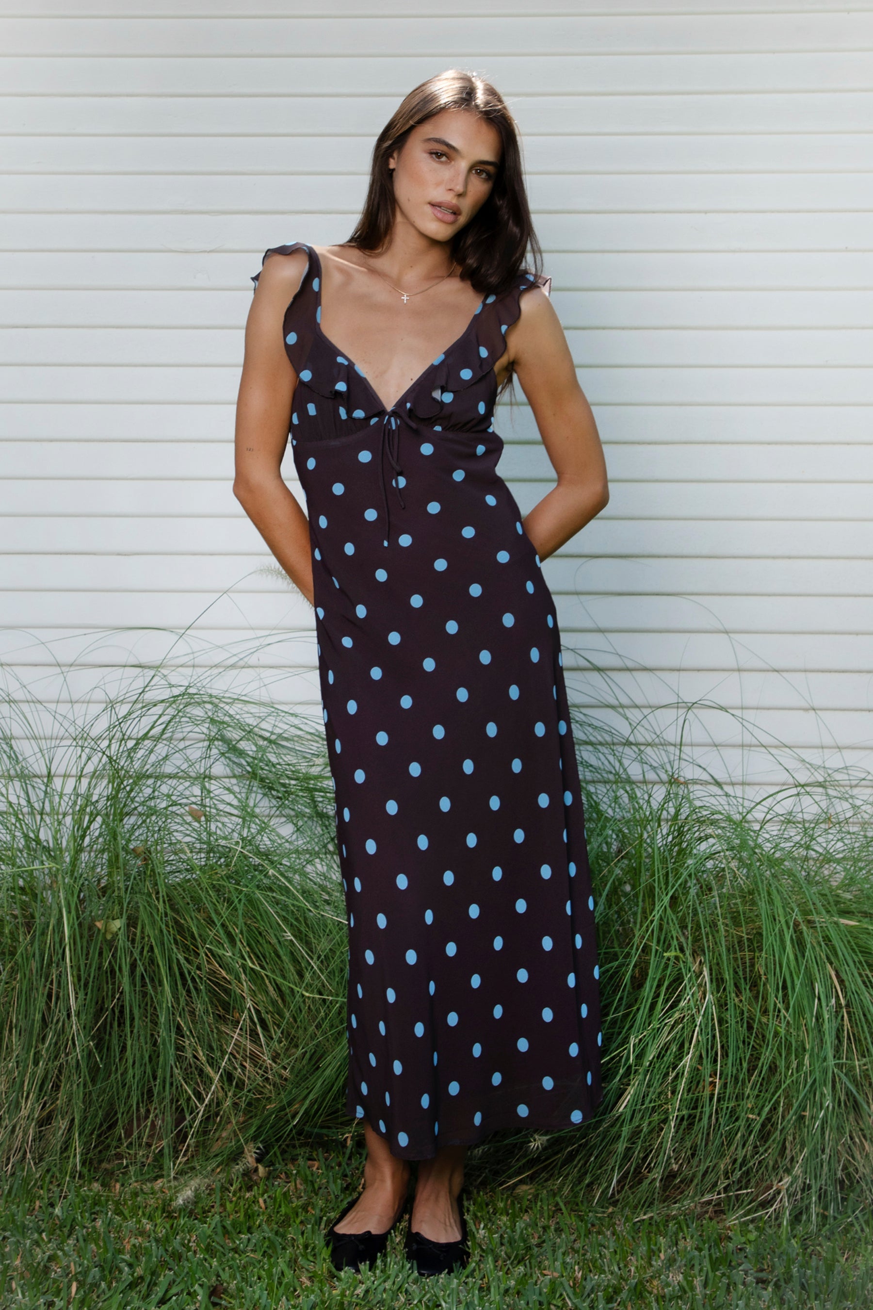 Woman wearing NOIRA ruffle chiffon polka dot maxi dress featuring flowy chiffon fabric, contrast polka dot print, deep V neckline with feminine ruffle trim, ruffled shoulder straps, adjustable front tie detail at the bust, fitted empire waist silhouette, low open back, adjustable shoulder straps, and soft stretch satin lining.