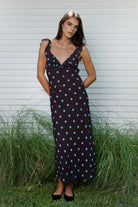 Woman wearing NOIRA ruffle chiffon polka dot maxi dress featuring flowy chiffon fabric, contrast polka dot print, deep V neckline with feminine ruffle trim, ruffled shoulder straps, adjustable front tie detail at the bust, fitted empire waist silhouette, low open back, adjustable shoulder straps, and soft stretch satin lining.