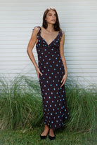Woman wearing NOIRA ruffle chiffon polka dot maxi dress featuring flowy chiffon fabric, contrast polka dot print, deep V neckline with feminine ruffle trim, ruffled shoulder straps, adjustable front tie detail at the bust, fitted empire waist silhouette, low open back, adjustable shoulder straps, and soft stretch satin lining.
