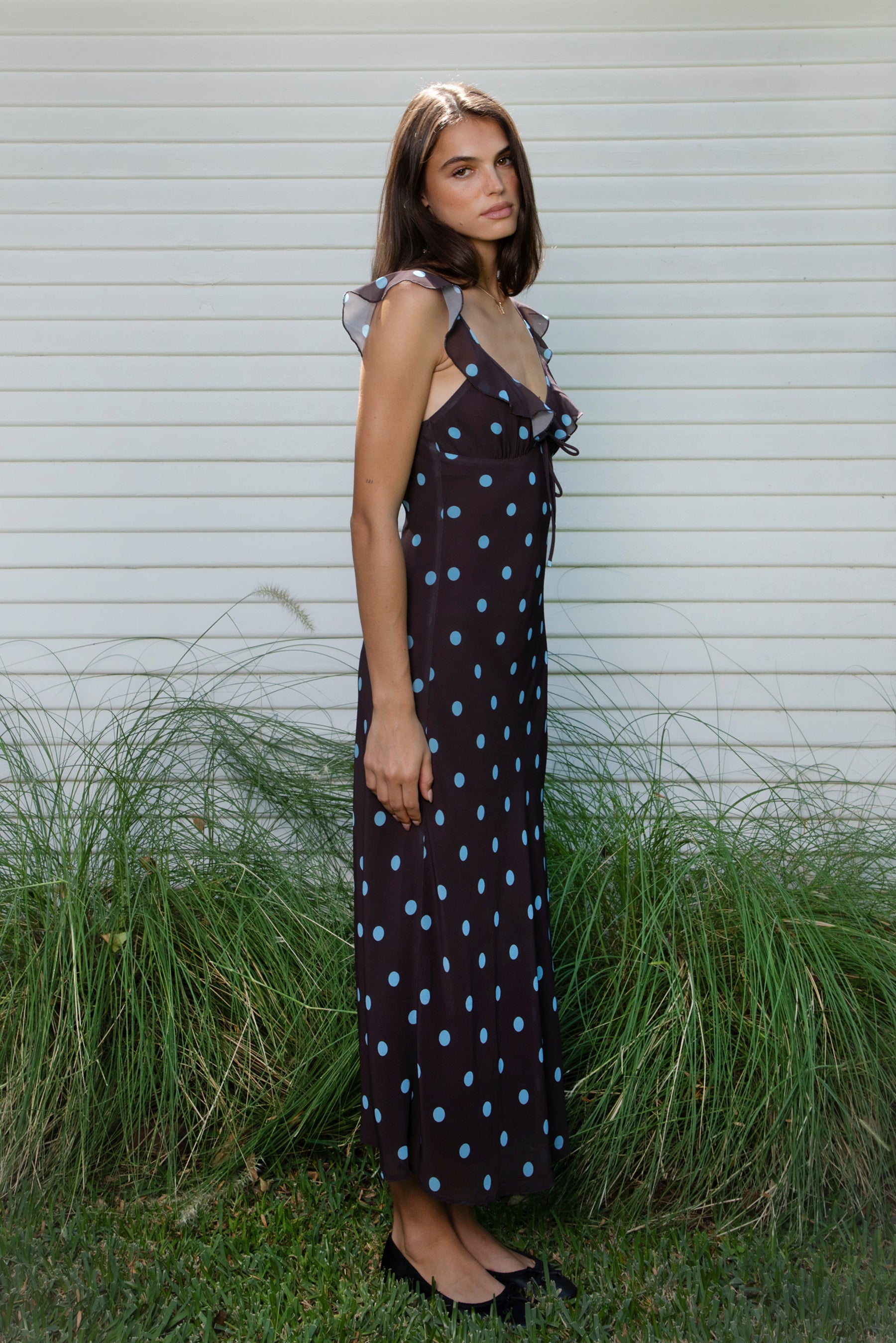 Woman wearing NOIRA ruffle chiffon polka dot maxi dress featuring flowy chiffon fabric, contrast polka dot print, deep V neckline with feminine ruffle trim, ruffled shoulder straps, adjustable front tie detail at the bust, fitted empire waist silhouette, low open back, adjustable shoulder straps, and soft stretch satin lining.