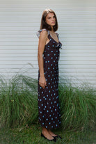 Woman wearing NOIRA ruffle chiffon polka dot maxi dress featuring flowy chiffon fabric, contrast polka dot print, deep V neckline with feminine ruffle trim, ruffled shoulder straps, adjustable front tie detail at the bust, fitted empire waist silhouette, low open back, adjustable shoulder straps, and soft stretch satin lining.