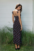 Woman wearing NOIRA ruffle chiffon polka dot maxi dress featuring flowy chiffon fabric, contrast polka dot print, deep V neckline with feminine ruffle trim, ruffled shoulder straps, adjustable front tie detail at the bust, fitted empire waist silhouette, low open back, adjustable shoulder straps, and soft stretch satin lining.