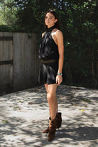 Woman wearing VESPER studded chiffon halter mini dress featuring flowy chiffon fabric, soft feminine drape, high halter neckline with self tie back closure, open back silhouette, gold studded geometric detailing along the neckline and waist, blouson bodice, gathered details, flared mini skirt, sheer chiffon overlay, and full lining.