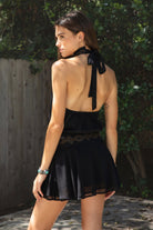 Woman wearing VESPER studded chiffon halter mini dress featuring flowy chiffon fabric, soft feminine drape, high halter neckline with self tie back closure, open back silhouette, gold studded geometric detailing along the neckline and waist, blouson bodice, gathered details, flared mini skirt, sheer chiffon overlay, and full lining.