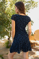 Women wearing TIC TOC COLLETE Floral Flocked Mesh Mini Dress in blue with V neckline, bow detail, and flirty godet skirt with greenery in the background. 