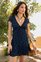 Women wearing TIC TOC COLLETE Floral Flocked Mesh Mini Dress in blue with V neckline, bow detail, and flirty godet skirt with greenery in the background. 