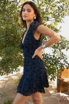 Women wearing TIC TOC COLLETE Floral Flocked Mesh Mini Dress in blue with V neckline, bow detail, and flirty godet skirt with greenery in the background. 