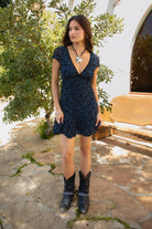 Women wearing TIC TOC COLLETE Floral Flocked Mesh Mini Dress in blue with V neckline, bow detail, and flirty godet skirt paired with black cowboy boots in a greenery in the background. 