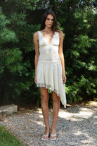Woman wearing FIORA flocked dot asymmetrical mini dress featuring soft flocked dot mesh, sleeveless silhouette, deep V neckline with gathered bust detail, ruched fitted waist, low V back neckline, asymmetrical handkerchief hem with extended side drape, and soft interlock jersey lining.