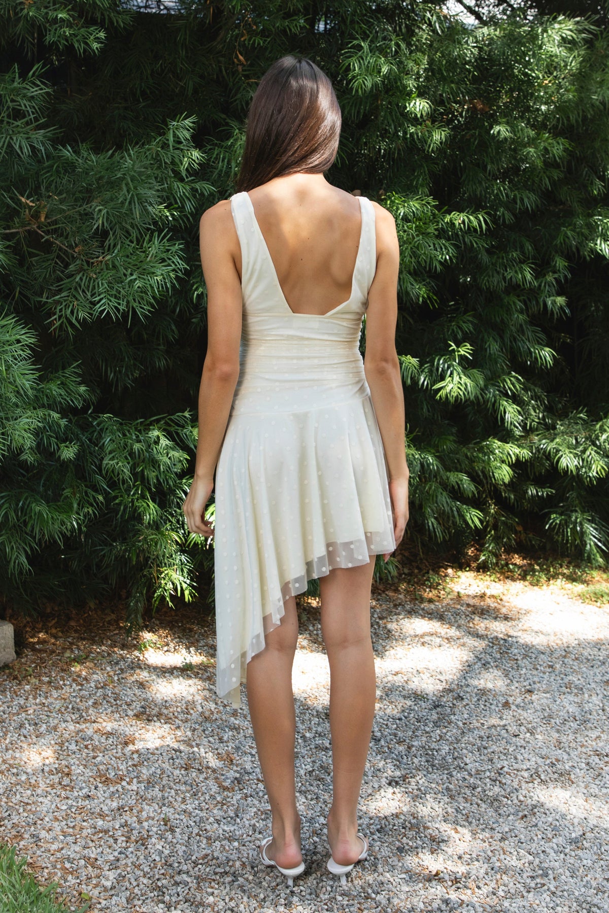 Woman wearing FIORA flocked dot asymmetrical mini dress featuring soft flocked dot mesh, sleeveless silhouette, deep V neckline with gathered bust detail, ruched fitted waist, low V back neckline, asymmetrical handkerchief hem with extended side drape, and soft interlock jersey lining.