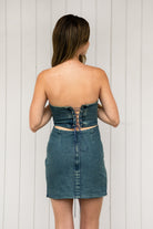 Woman wearing ARIZONA denim western stitch mini dress featuring vintage washed denim, bold western inspired contrast stitching, strapless corset style bodice, lace up back with adjustable closure, subtle back cutout, fitted waist, and a soft stretch mini length silhouette.