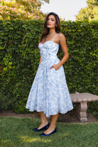 Woman in a light blue floral dress standing outdoors with greenery in the background