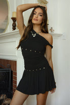 Women wearing TIC TOC AZALEA One Shoulder Mini Dress in black with twisted strap, ruching, studded detail, and flirty hemline in doors near a fire place