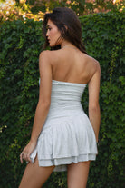 Woman wearing TIC TOC LOREA Flirty Hem Tube Romper Mini Dress in [Color] with ruched bodice, tiered skirt, and built-in shorts standing against a green hedge.