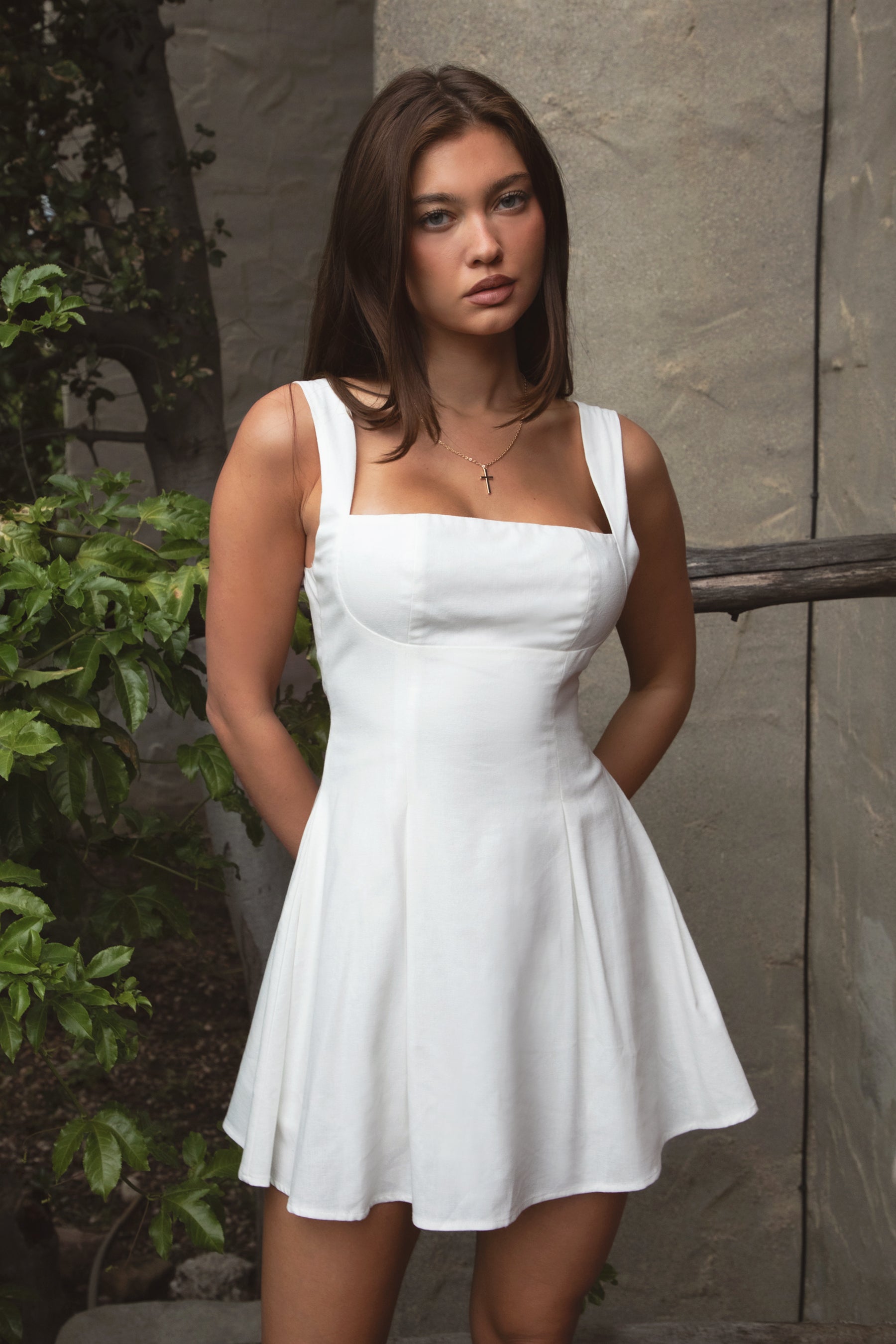 Woman wearing TIC TOC DH31916-ZENAYA ZENAYA mini dress featuring a fit-and-flare mini silhouette, clean square neckline, adjustable wide shoulder straps, shaped bust seams, and a bow tie open back detail, with soft linen-blend drape and lightweight structure styled for a summer-ready brunch or garden party look.