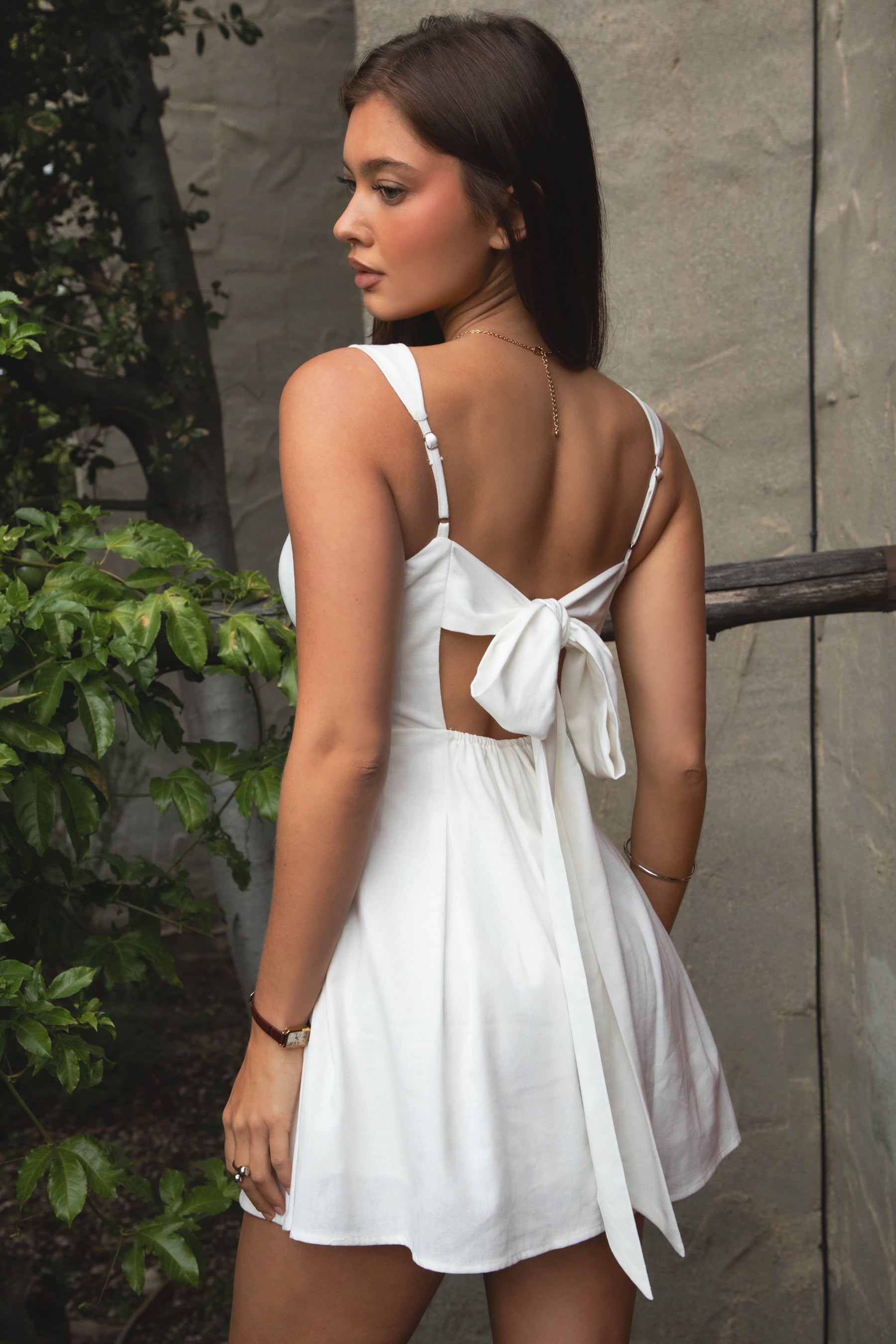 Woman wearing TIC TOC DH31916-ZENAYA ZENAYA mini dress featuring a fit-and-flare mini silhouette, clean square neckline, adjustable wide shoulder straps, shaped bust seams, and a bow tie open back detail, with soft linen-blend drape and lightweight structure styled for a summer-ready brunch or garden party look.