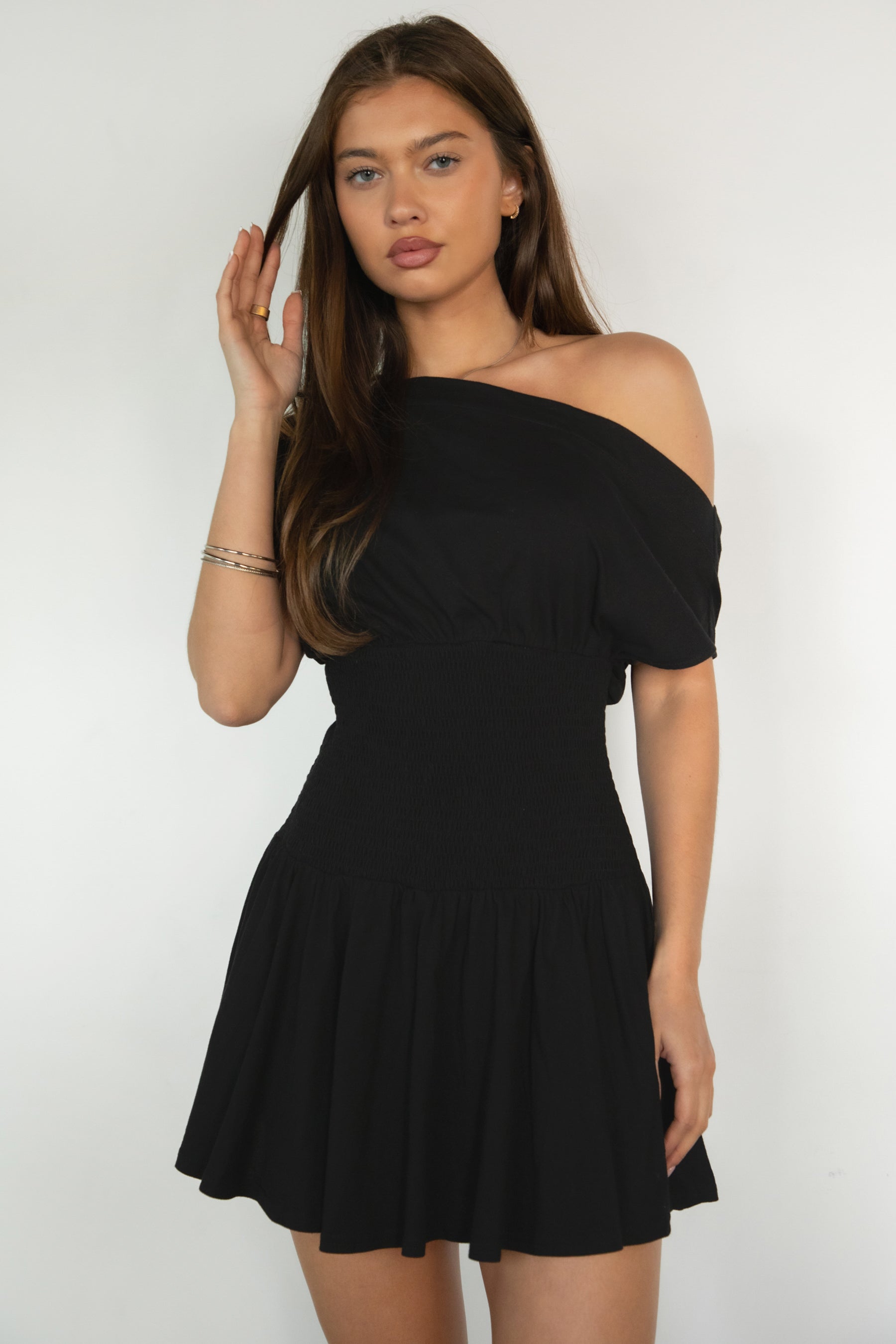 Woman wearing TIC TOC DH31921-ASHA ASHA asymmetry shirt sleeve mini dress featuring an asymmetrical one-shoulder shirt sleeve, soft cotton jersey fabrication, slouchy off-the-shoulder neckline, smocked fitted bodice, and a gathered swingy flared mini skirt, styled simply with sandals for an easy summer daytime look.