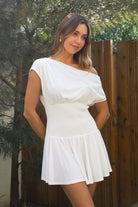 Woman wearing TIC TOC DH31921-ASHA ASHA asymmetry shirt sleeve mini dress featuring an asymmetrical one-shoulder shirt sleeve, soft cotton jersey fabrication, slouchy off-the-shoulder neckline, smocked fitted bodice, and a gathered swingy flared mini skirt, styled simply with sandals for an easy summer daytime look.