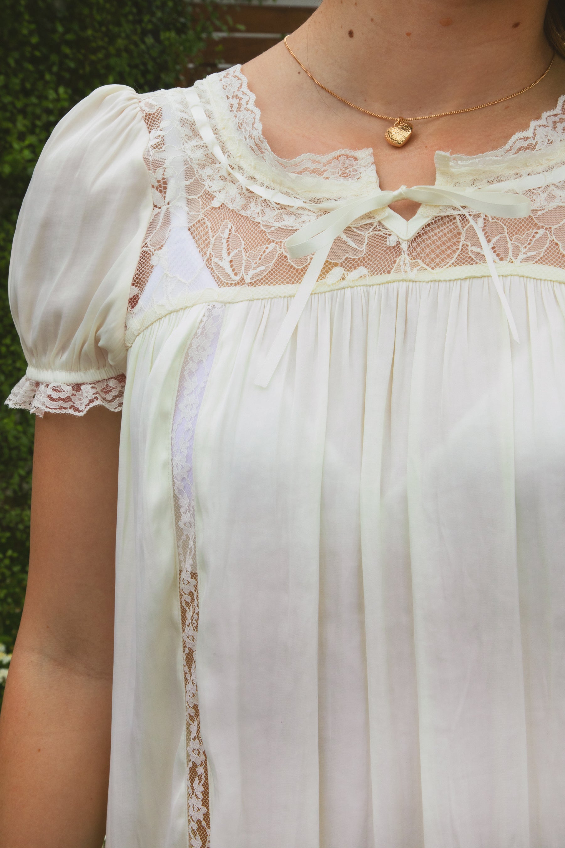 Woman wearing EVANGELIN sheer satin short sleeve mini dress featuring a romantic scoop neckline with sheer lace trim, soft satin babydoll silhouette, puff sleeves with elastic lace cuffs, front self tie bow, lace contrast panels, and ruffle hem finish for bridal and romantic occasion styling.