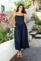 Woman wearing MAYREN smocked bow back denim Tencel maxi dress featuring soft denim Tencel fabric, comfortable stretch, straight neckline, adjustable spaghetti straps, smocked bodice, statement open back with oversized self tie bow detail, side seam pockets, and flowing maxi length skirt.