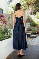 Woman wearing MAYREN smocked bow back denim Tencel maxi dress featuring soft denim Tencel fabric, comfortable stretch, straight neckline, adjustable spaghetti straps, smocked bodice, statement open back with oversized self tie bow detail, side seam pockets, and flowing maxi length skirt.
