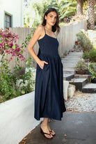 Woman wearing MAYREN smocked bow back denim Tencel maxi dress featuring soft denim Tencel fabric, comfortable stretch, straight neckline, adjustable spaghetti straps, smocked bodice, statement open back with oversized self tie bow detail, side seam pockets, and flowing maxi length skirt.