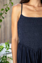 Woman wearing MAYREN smocked bow back denim Tencel maxi dress featuring soft denim Tencel fabric, comfortable stretch, straight neckline, adjustable spaghetti straps, smocked bodice, statement open back with oversized self tie bow detail, side seam pockets, and flowing maxi length skirt.