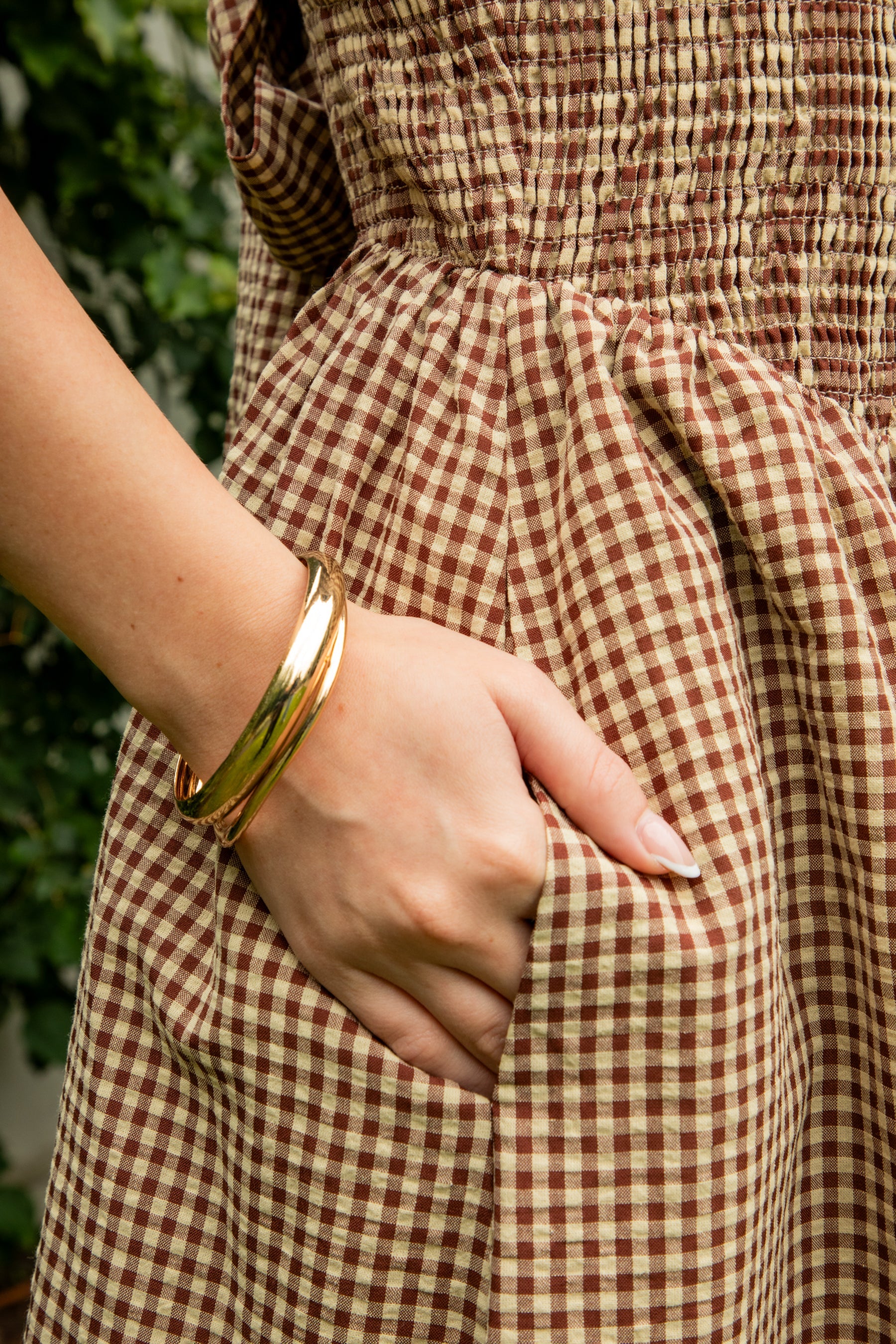 Woman wearing MAYREN gingham smocked midi dress featuring a classic gingham check pattern, fully smocked bodice, adjustable spaghetti straps, square neckline, open back with adjustable tie detail, subtle cutout accent, side seam pockets, and a flowing gathered midi skirt.
