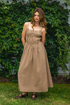 Woman wearing MAYREN gingham smocked midi dress featuring a classic gingham check pattern, fully smocked bodice, adjustable spaghetti straps, square neckline, open back with adjustable tie detail, subtle cutout accent, side seam pockets, and a flowing gathered midi skirt.