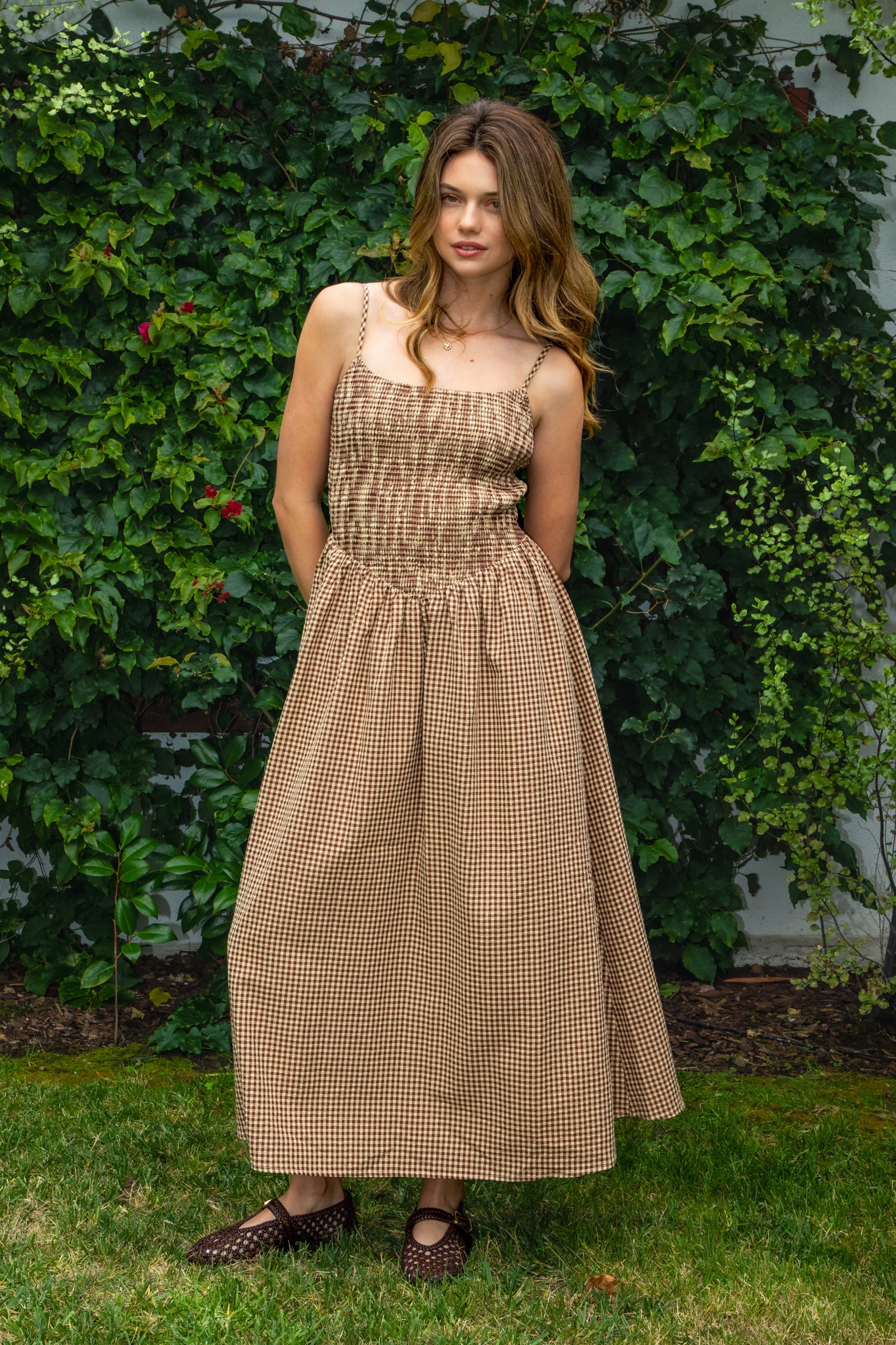 Woman wearing MAYREN gingham smocked midi dress featuring a classic gingham check pattern, fully smocked bodice, adjustable spaghetti straps, square neckline, open back with adjustable tie detail, subtle cutout accent, side seam pockets, and a flowing gathered midi skirt.