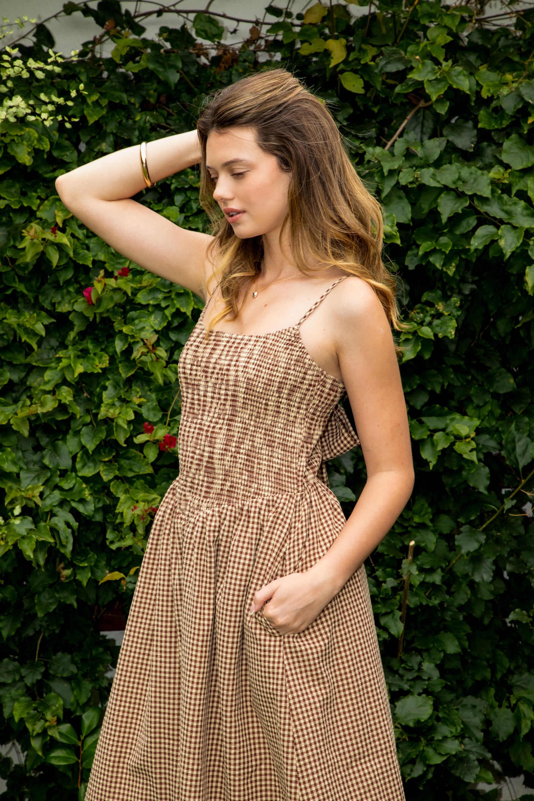 Woman wearing MAYREN gingham smocked midi dress featuring a classic gingham check pattern, fully smocked bodice, adjustable spaghetti straps, square neckline, open back with adjustable tie detail, subtle cutout accent, side seam pockets, and a flowing gathered midi skirt.