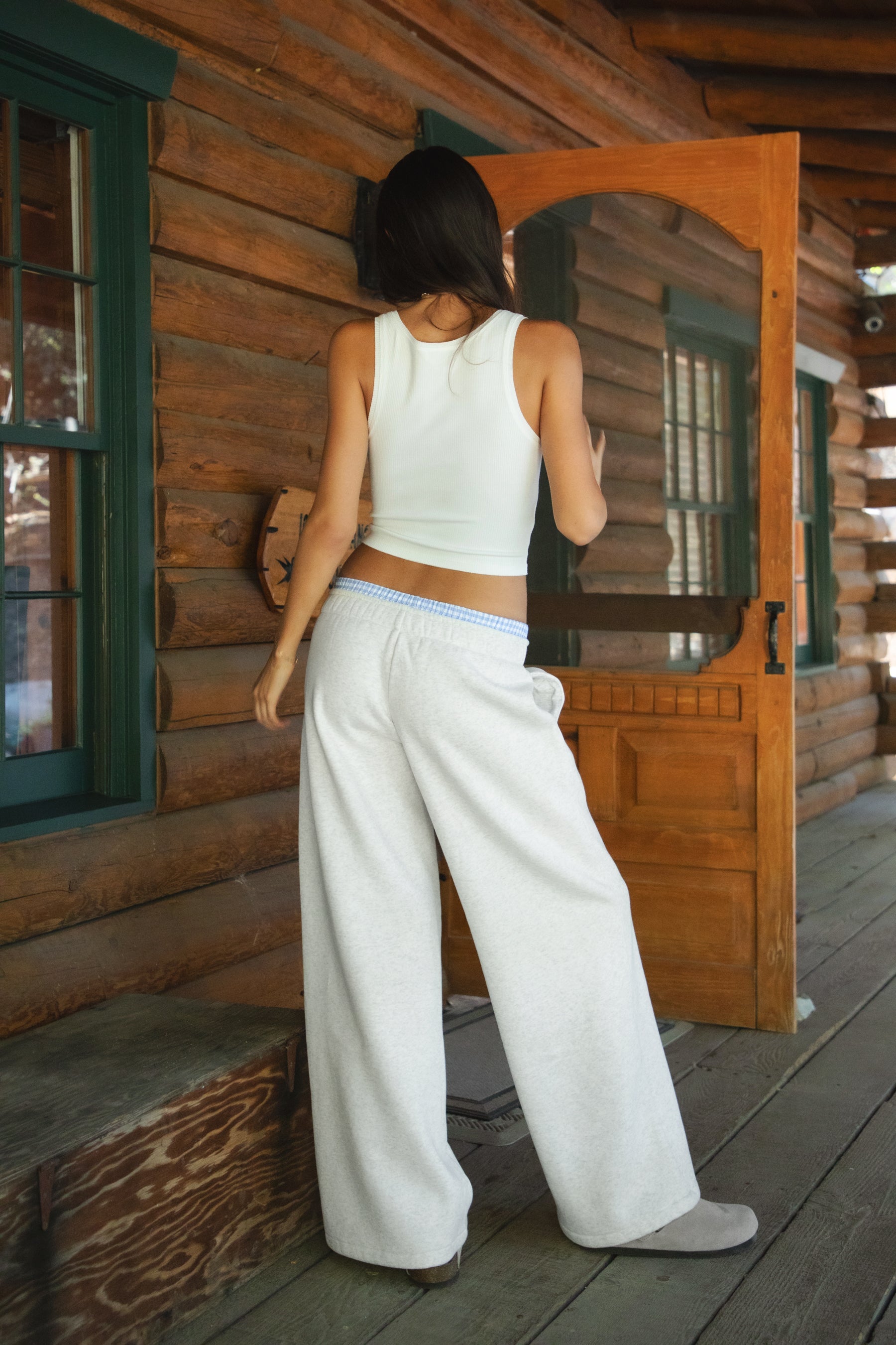 Woman wearing heather white TIC TOC CASSE Fleece Sweatpants with built-in gingham boxer, back view with a matching rib tank top in a cabin. 