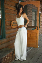 Woman wearing heather white TIC TOC CASSE Fleece Sweatpants with built-in gingham boxer, side view with a matching rib tank top in a cabin. 
