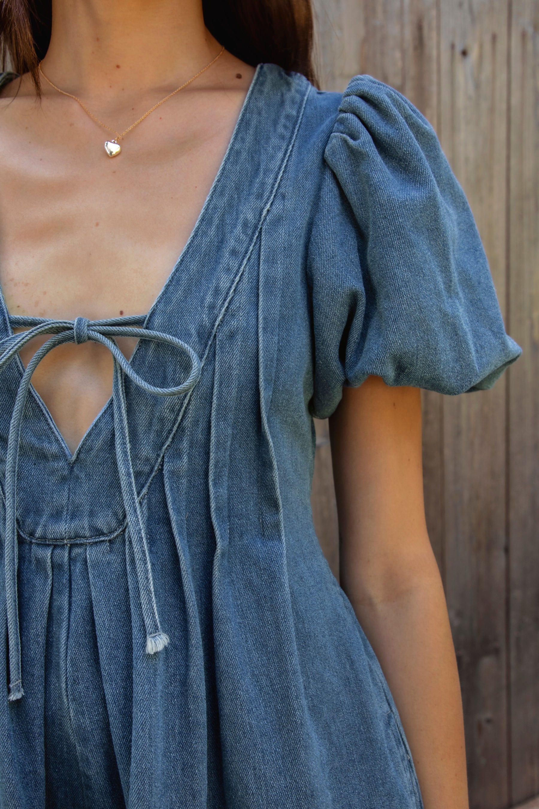 Woman wearing DELANEY soft denim puff sleeve denim romper featuring soft denim fabric, comfortable structured feel, deep V neckline, adjustable front tie detail, statement puff sleeves with gathered volume, pleated bodice, relaxed fit silhouette, and hidden side zipper closure.