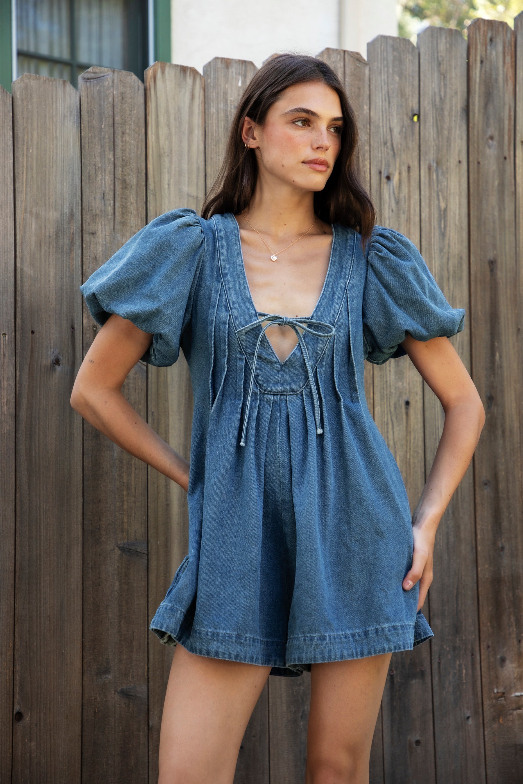 Woman wearing DELANEY soft denim puff sleeve denim romper featuring soft denim fabric, comfortable structured feel, deep V neckline, adjustable front tie detail, statement puff sleeves with gathered volume, pleated bodice, relaxed fit silhouette, and hidden side zipper closure.